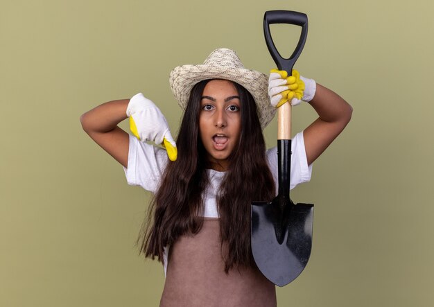 Chica joven jardinero en delantal y sombrero de verano con guantes de trabajo sosteniendo la pala sorprendida pointign con el dedo índice hacia abajo de pie sobre la pared verde