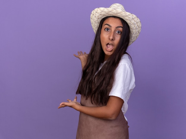 Chica joven jardinero en delantal y sombrero de verano presentando algo hacia atrás con los brazos de pie sobre la pared púrpura