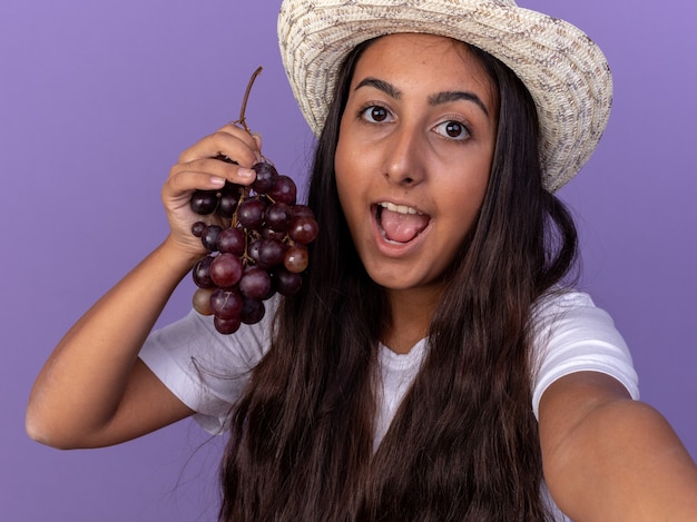 Chica joven jardinero en delantal y sombrero de verano con racimo de uva sonriendo con cara feliz de pie sobre la pared púrpura