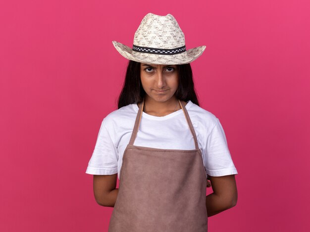 Chica joven jardinero en delantal y sombrero de verano con rostro serio parado sobre pared rosa