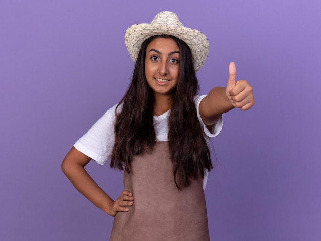 Chica joven jardinero en delantal y sombrero de verano sonriendo alegremente mostrando los pulgares para arriba de pie sobre la pared púrpura