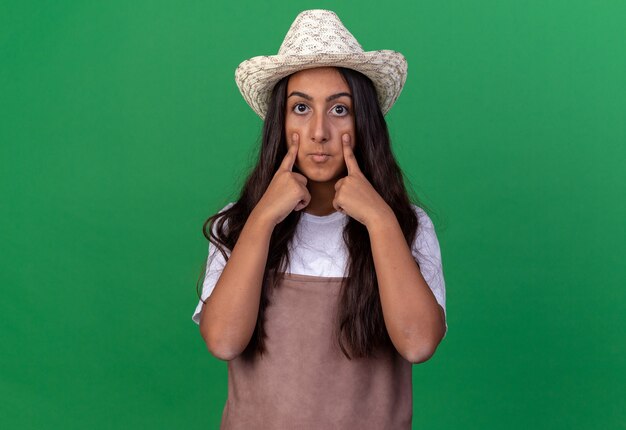 Chica joven jardinero en delantal y sombrero de verano sorprendido señalando con el dedo índice a sus ojos de pie sobre la pared verde