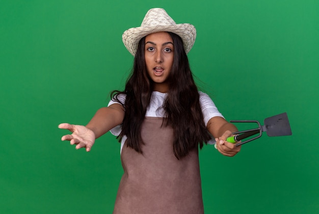 Foto gratuita chica joven jardinero en delantal y sombrero de verano sosteniendo azadón amplia apertura manos sorprendidas de pie sobre la pared verde