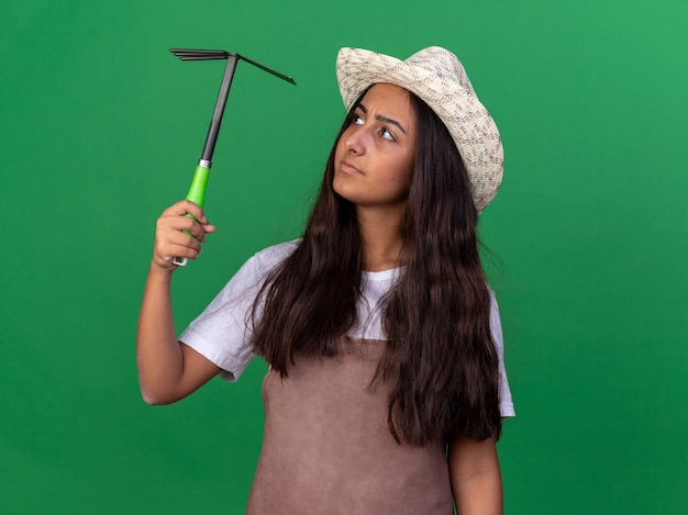 Chica joven jardinero en delantal y sombrero de verano sosteniendo azadón mirándolo intrigado de pie sobre la pared verde