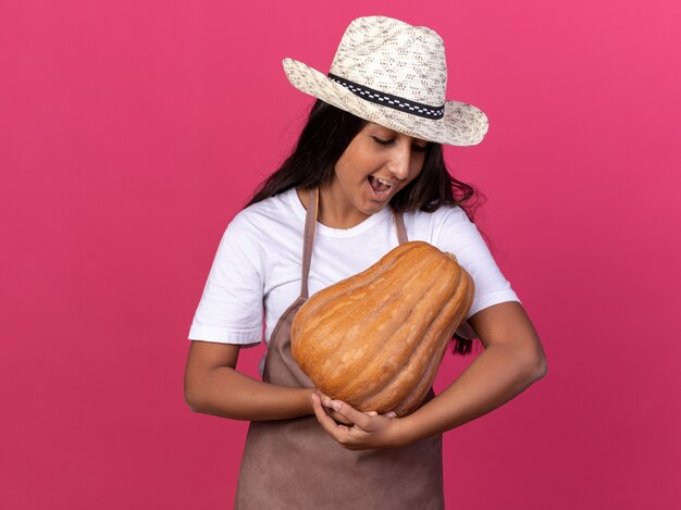 Chica joven jardinero en delantal y sombrero de verano sosteniendo calabaza mirándola con una sonrisa en la cara feliz y alegre de pie sobre la pared rosa