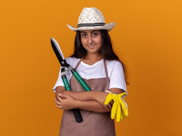 Chica joven jardinero en delantal y sombrero de verano sosteniendo cortasetos y guantes de trabajo con una sonrisa en la cara de pie sobre la pared naranja