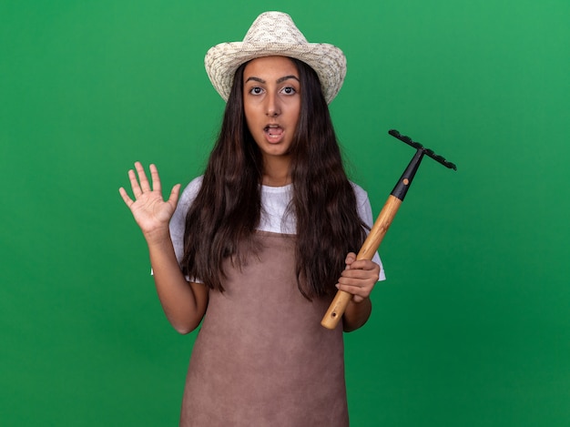 Chica joven jardinero en delantal y sombrero de verano sosteniendo mini rastrillo sorprendido y sorprendido levantando el brazo sobre la pared verde