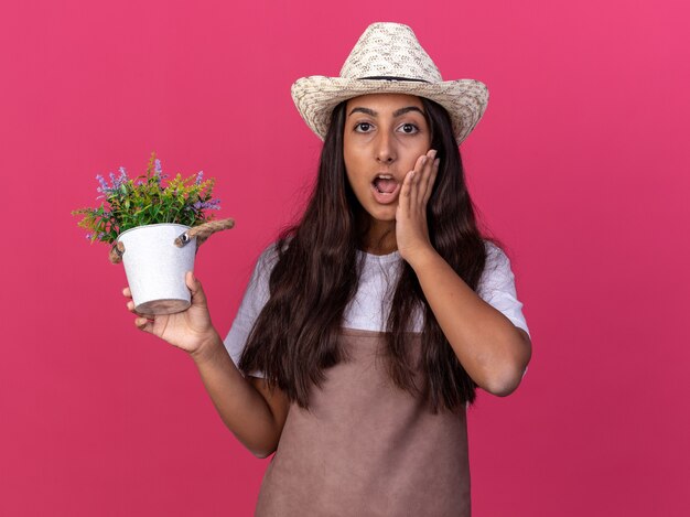 Chica joven jardinero en delantal y sombrero de verano sosteniendo planta en maceta asombrado y sorprendido de pie sobre la pared rosa