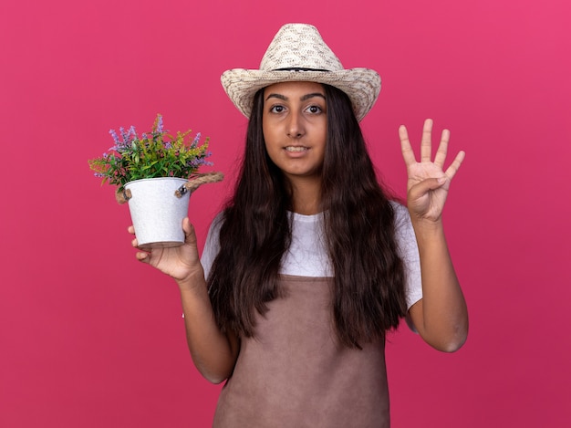 Chica joven jardinero en delantal y sombrero de verano sosteniendo una planta en maceta sonriendo mostrando el número cuatro de pie sobre la pared rosa
