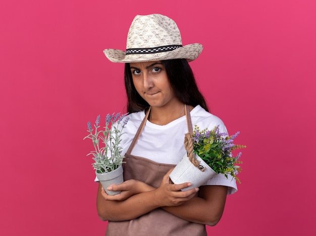 Chica joven jardinero en delantal y sombrero de verano sosteniendo plantas en macetas con cara enojada de pie sobre la pared rosa