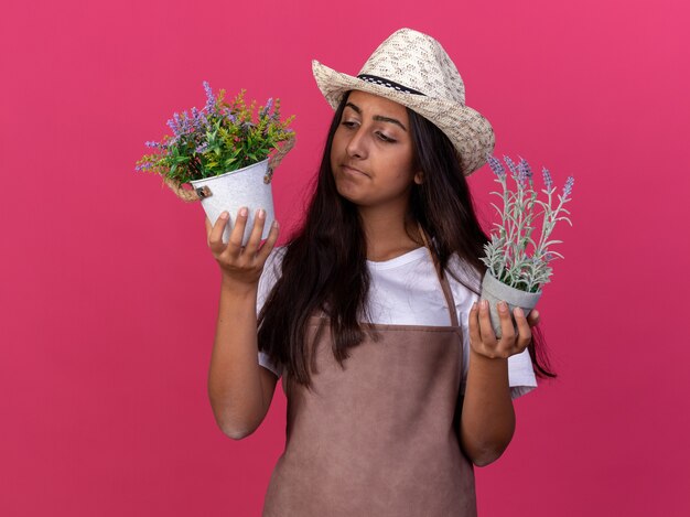 Chica joven jardinero en delantal y sombrero de verano sosteniendo plantas en macetas mirándolas confundirse de pie sobre la pared rosa