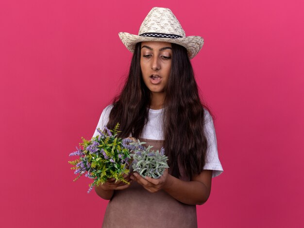 Chica joven jardinero en delantal y sombrero de verano sosteniendo plantas en macetas mirándolos sorprendido de pie sobre la pared rosa