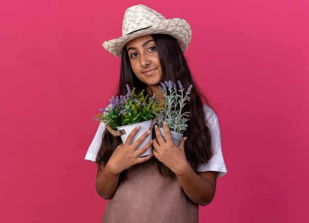 Chica joven jardinero en delantal y sombrero de verano sosteniendo plantas en macetas sonriendo con cara feliz de pie sobre la pared rosa