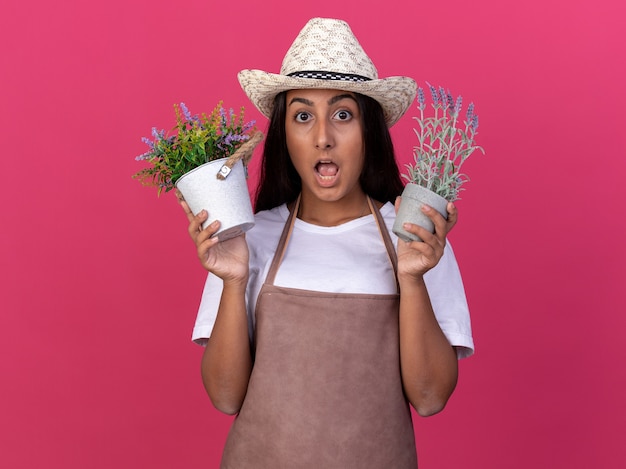 Chica joven jardinero en delantal y sombrero de verano sosteniendo plantas en macetas sorprendió de pie sobre la pared rosa