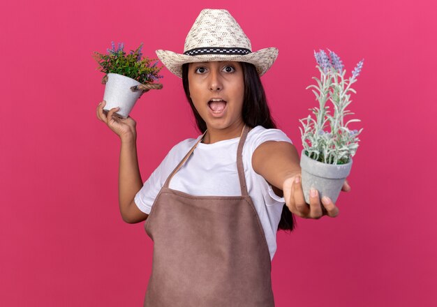 Chica joven jardinero en delantal y sombrero de verano sosteniendo plantas en macetas sorprendió de pie sobre la pared rosa