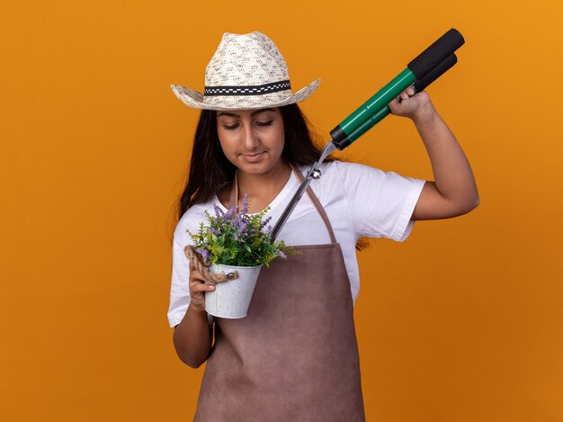 Chica joven jardinero en delantal y sombrero de verano sosteniendo podadoras de setos y planta en maceta loking seguro sonriente de pie sobre pared naranja