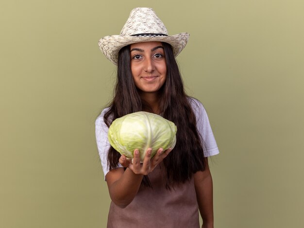 Chica joven jardinero en delantal y sombrero de verano sosteniendo repollo con sonrisa en la cara de pie sobre la pared verde