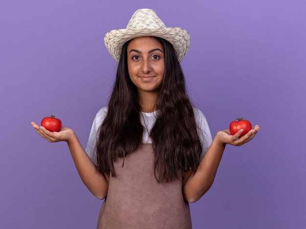 Chica joven jardinero en delantal y sombrero de verano sosteniendo tomates frescos con una sonrisa en la cara de pie sobre la pared púrpura