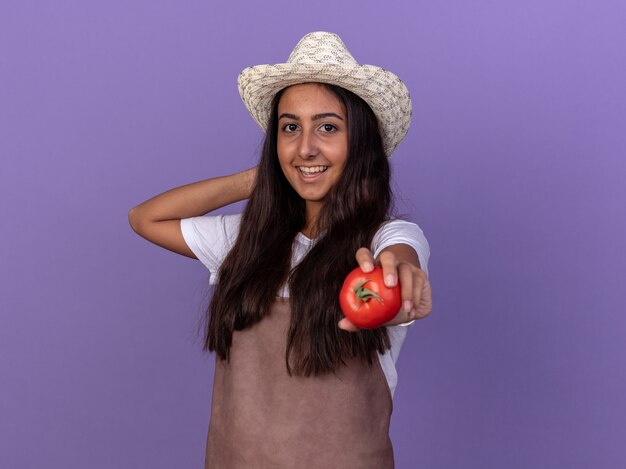 Chica joven jardinero en delantal y sombrero de verano sosteniendo tomates frescos con una sonrisa en la cara de pie sobre la pared púrpura