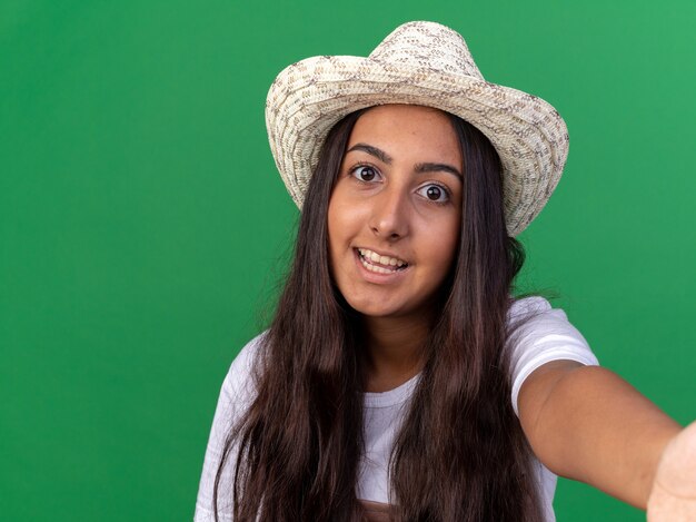 Chica joven jardinero feliz en delantal y sombrero de verano feliz y alegre sonriente de pie sobre la pared verde