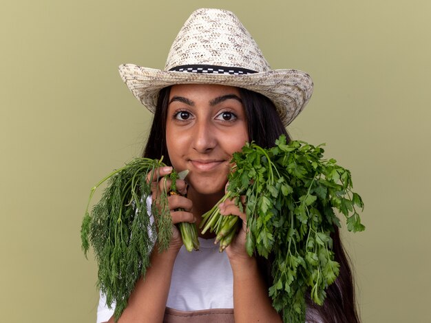 Chica joven jardinero feliz en delantal y sombrero de verano con hierbas frescas con una sonrisa en la cara de pie sobre la pared verde