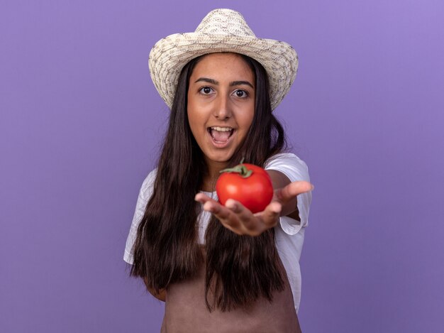 Chica joven jardinero feliz en delantal y sombrero de verano mostrando tomates frescos con una sonrisa en la cara de pie sobre la pared púrpura