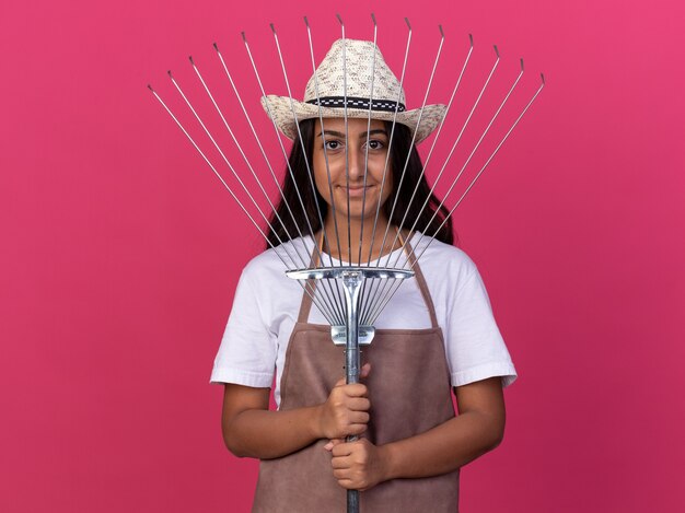 Chica joven jardinero feliz en delantal y sombrero de verano con rastrillo con una sonrisa en la cara de pie sobre la pared rosa