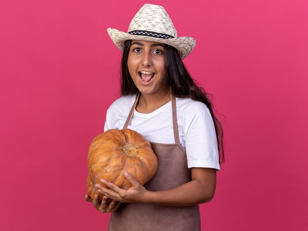 Chica joven jardinero feliz y sorprendida en delantal y sombrero de verano con calabaza con sonrisa en la cara de pie sobre la pared rosa