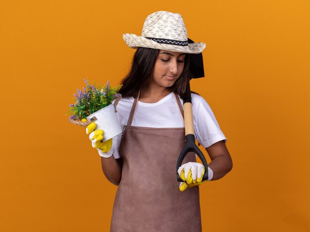 Chica joven jardinero sosteniendo una planta en maceta y una pala mirando confiado de pie sobre la pared naranja