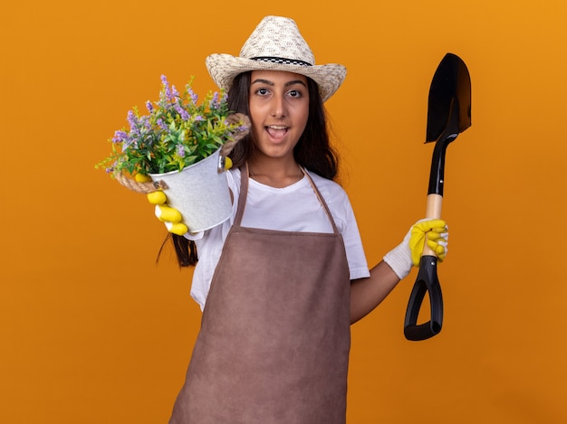 Chica joven jardinero sosteniendo una planta en maceta y una pala sonriendo alegremente de pie sobre la pared naranja