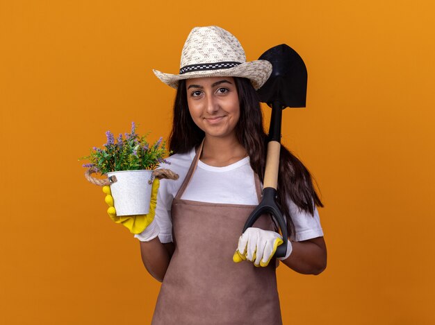 Chica joven jardinero sosteniendo una planta en maceta y una pala con una sonrisa en la cara de pie sobre la pared naranja