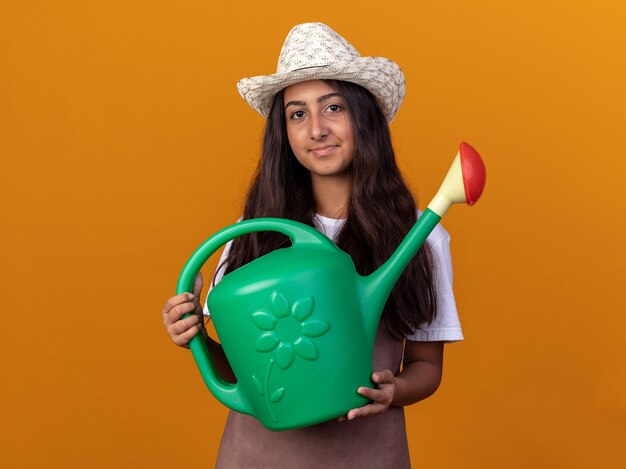 Chica joven jardinero sosteniendo regadera con una sonrisa en la cara de pie sobre la pared naranja