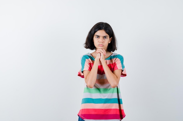 Chica joven juntando las manos debajo de la barbilla en camiseta a rayas de colores y mirando seria. vista frontal.