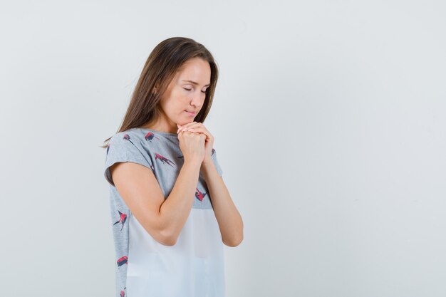 Chica joven juntando las manos en gesto de oración en camiseta y mirando esperanzado, vista frontal.