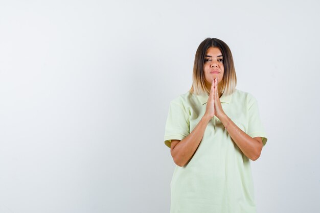 Chica joven juntando las manos en posición de oración en camiseta y mirando serio, vista frontal.