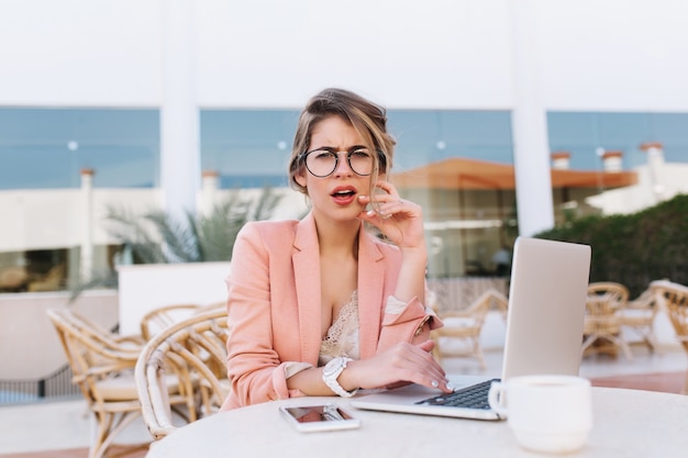 Chica joven con laptop en café de la calle con expresión facial disgustada, mirada molesta o insatisfecha. Vistiendo elegante chaqueta rosa, gafas, relojes blancos.