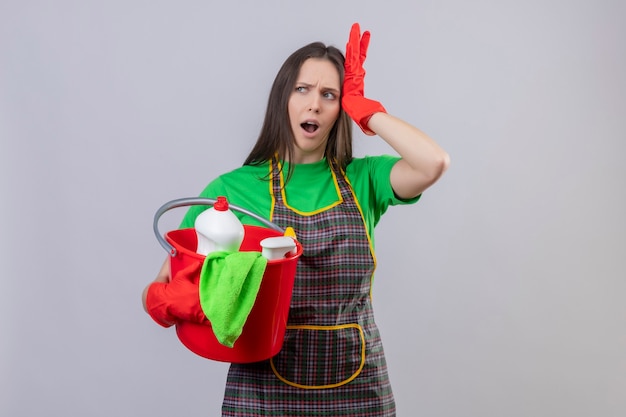 Chica joven de limpieza descontenta con uniforme en guantes rojos sosteniendo herramientas de limpieza puso su mano en la cabeza en la pared blanca aislada