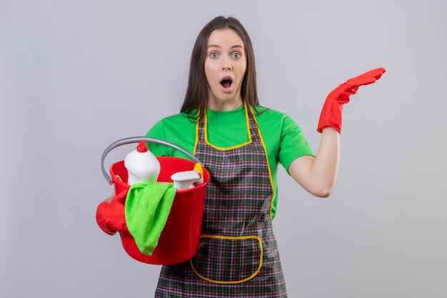 Chica joven de limpieza sorprendida con uniforme en guantes rojos sosteniendo herramientas de limpieza levantando la mano sobre fondo blanco aislado