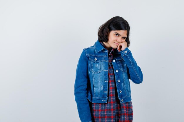 Chica joven con la mano apoyada en la mejilla en camisa a cuadros y chaqueta de jean y luciendo linda, vista frontal.