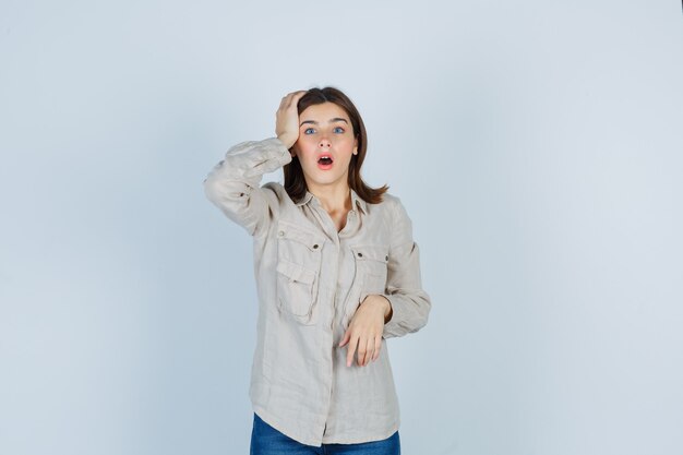 Chica joven con la mano en la cabeza, manteniendo la boca abierta en camisa beige, jeans y mirando sorprendido, vista frontal.