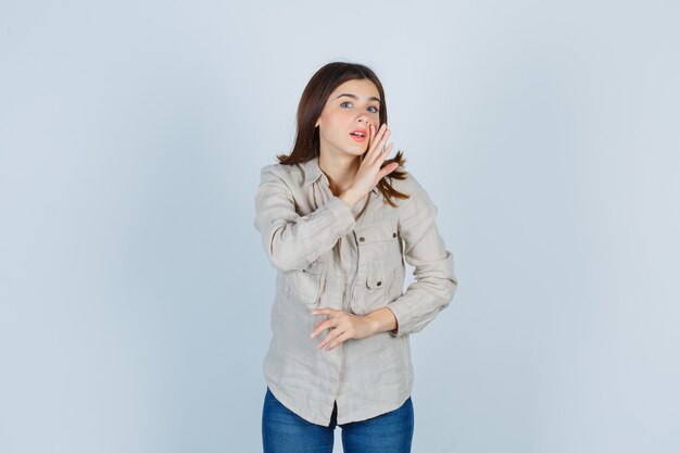 Chica joven con la mano cerca de la boca como diciendo un secreto en camisa beige, jeans y mirando enfocado, vista frontal.