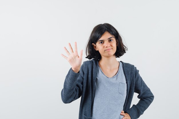 Chica joven con la mano en la cintura, levantando la mano saludando a alguien con una camiseta gris claro y una sudadera con capucha gris oscuro y con un aspecto lindo.