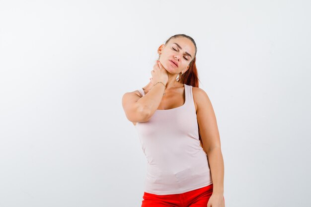 Chica joven con la mano en el cuello, con dolor de cuello en top beige y pantalones rojos y luciendo exhausta. vista frontal.