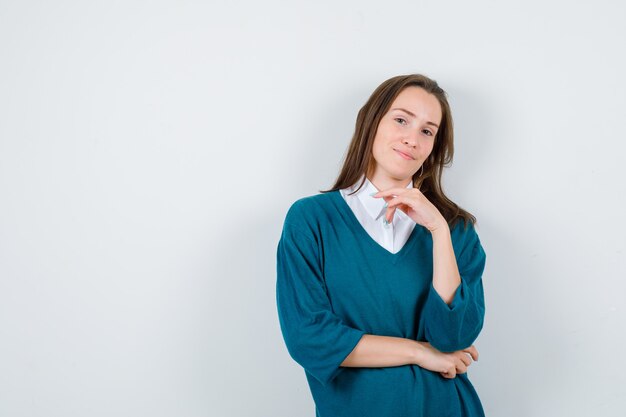 Chica joven con la mano debajo de la barbilla en un suéter con cuello en v, camisa y una linda vista frontal.