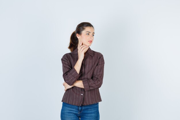 Chica joven con la mano en la papada, mirando al lado derecho con camisa a rayas, jeans y mirando enfocado, vista frontal.