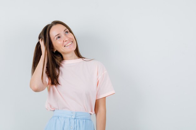 Chica joven con la mano en el pelo en camiseta, falda y mirando esperanzado, vista frontal.