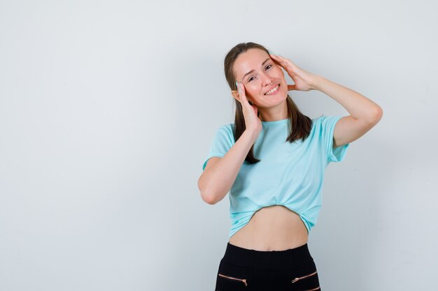 Chica joven con las manos en la cara en camiseta turquesa, pantalones y mirando feliz, vista frontal.