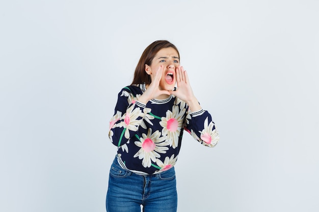 Chica joven con las manos cerca de la boca como llamando a alguien en suéter floral, jeans y gritando, vista frontal.