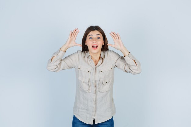 Chica joven con las manos cerca de la cabeza, manteniendo la boca abierta en camisa beige, jeans y mirando sorprendido. vista frontal.