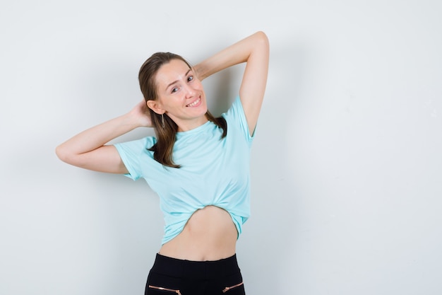 Foto gratuita chica joven con las manos detrás de la cabeza en camiseta turquesa, pantalones y mirando alegre, vista frontal.